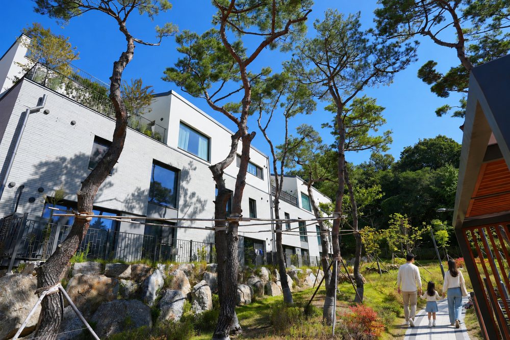 라피아노 용인 단지 산책길을 걸어 들어가는 가족의 뒷모습을 그린 연출 장면, 소나무와 흰 3층 건물